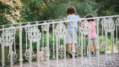 Two young children walk across an ornate white metal bridge with decorative railings, looking over the side toward water below. Trees and greenery fill the background.